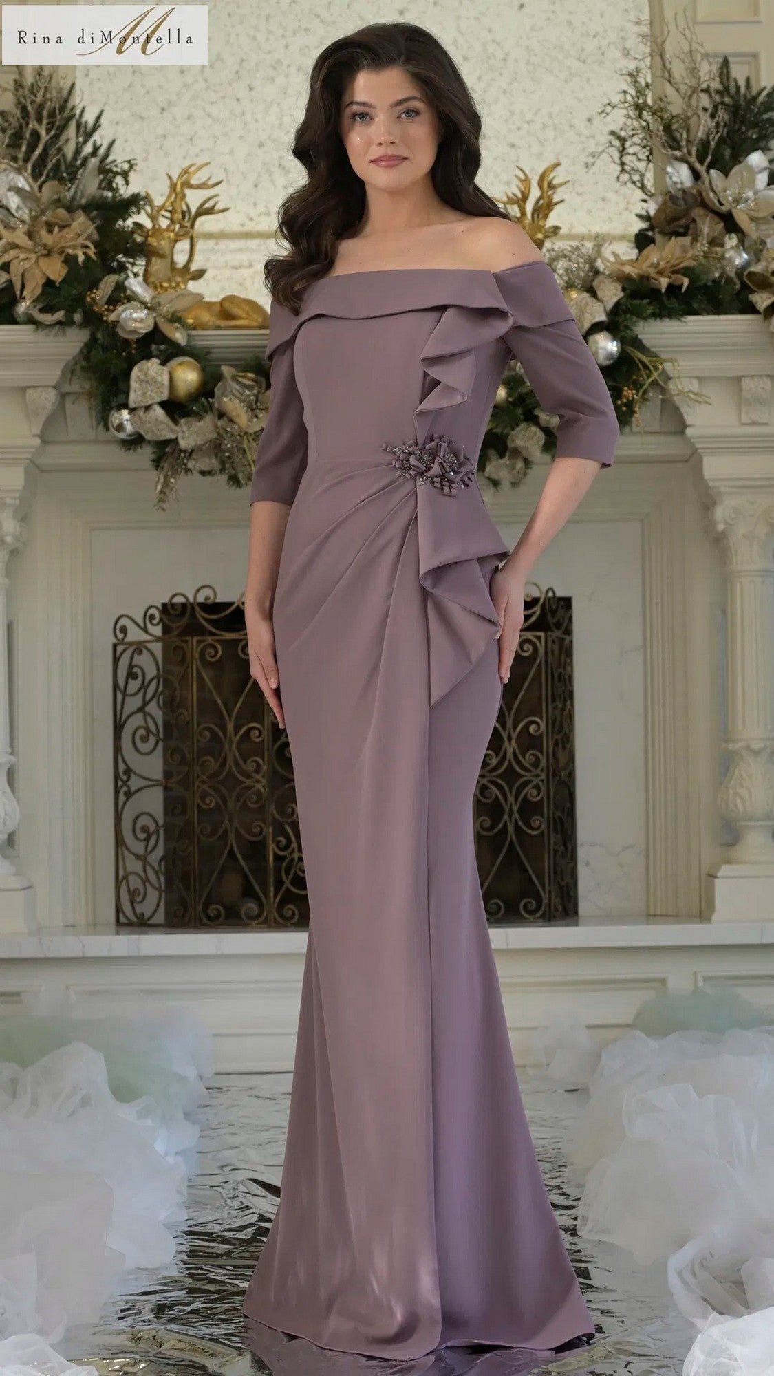 A woman stands indoors wearing the Rina Di Montella RD3202 long formal dress in Victorian Lilac, featuring off-the-shoulder ruffle details and floral embellishments at the waist, with decorative elements in the background.